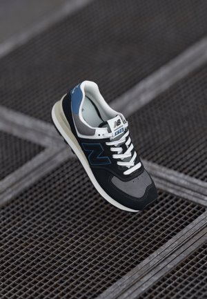 Pojedynczy but sportowy New Balance 574 w kolorach czarnym, szarym, niebieskim i białym unoszący się nad metalową kratą.