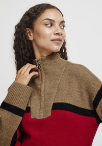 Strickpullover mit einem tannfarbenen Körper, roten und schwarzen Streifenakzenten, hohem Kragen und halbem Reißverschluss. Weiche Textur und lässige Passform.