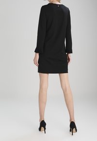 Svart klänning med långa ärmar som har volangdetaljer vid ärmsluten och en nyckelhålsknapp på baksidan, i kombination med svarta högklackade skor.