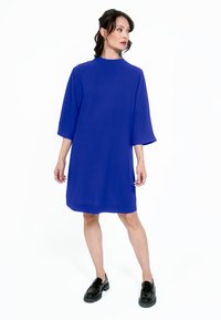 Kungligt blå bomullsklänning med hög hals, vida ärmar och lös passform, kombinerad med svarta loafers med platåsula. Mjukt, slätt tyg utan struktur.