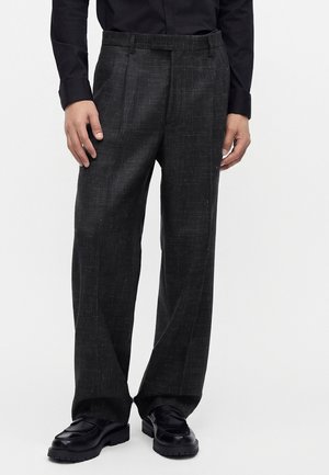 Homme portant un pantalon large noir texturé, une chemise noire et des mocassins en cuir noir, debout devant un fond uni.