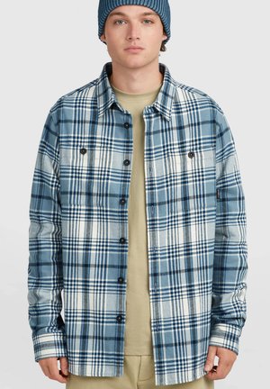 Junger Mann, der eine blaue Mütze und ein blau-weiß kariertes Hemd über einem beigen T-Shirt trägt, steht vor einem schlichten Hintergrund.