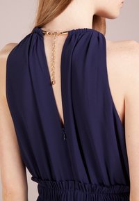 Mörkblå ärmlös klänning med en nyckelhålsspringa bak, samlad halsringning och guldig kedjedetalj. Har en åtsittande midja och dragkedjestängning.