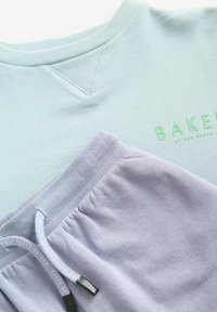 Felpa in cotone azzurro chiaro con texture a coste e logo verde "BAKER", insieme a pantaloni corti lilla con vita elastica e cordoncino.
