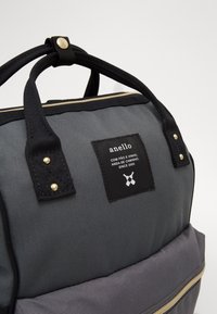 Szaro-czarna torba plecakowa z podwójnymi czarnymi uchwytami, przednią kieszenią oraz czarną etykietą z białym tekstem i skrzyżowanymi ikonami butelek.