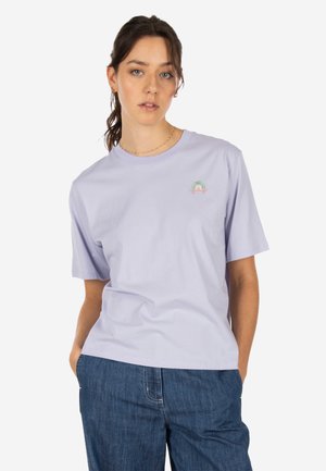 Junge Frau mit zurückgebundenen braunen Haaren, die ein lavendelfarbenes T-Shirt mit einem kleinen Regenbogenaufnäher und blaue Jeans trägt, die Hände in den Taschen.