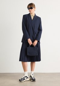Námořnický blazer s přiléhavým střihem a límci, spárovaný s plissé námořnickou sukní. Doplňky zahrnují malou texturovanou tašku a sportovní tenisky.