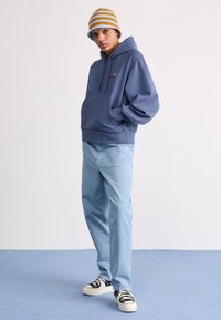 Haut à capuche bleu clair avec poche frontale, logo brodé. Pantalon bleu clair, baskets noir et blanc, et bonnet rayé orange et bleu.