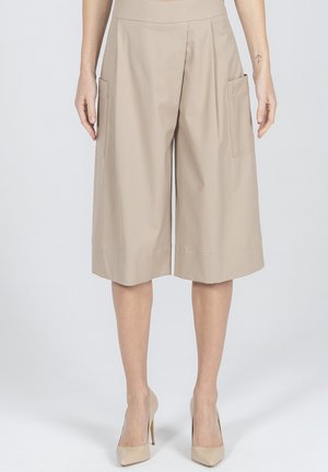 Beżowe, szerokie spodenki do kolan z dużymi kieszeniami po bokach, noszone z beżowymi szpilkami, na osobie stojącej na jednolitym tle.