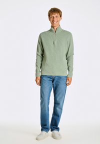 Ljusgröna ribbstickade långärmade tröja med halvvitt krage och dragkedja, kombinerad med blå jeans och vita sneakers, presenterad mot en vit bakgrund.