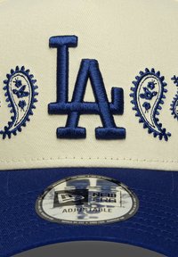 Cap mit einer cremefarbenen Vorderseite, blauen Schirm und dunkelblauem gesticktem "LA"-Logo. Hat Paisley-Muster an den Seiten. Verstellbarer Riemen hinten.
