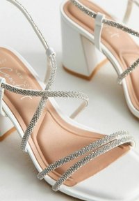 Sandali con tacco bianco e cinturini incrociati in strass argentati, con una soletta beige, caratterizzati da un tacco quadrato e una texture liscia.