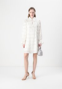 Robe blanche avec des détails en franges texturées, manches longues avec des poignets volantés, col, et un petit sac à main gris. Sandales avec des talons transparents.