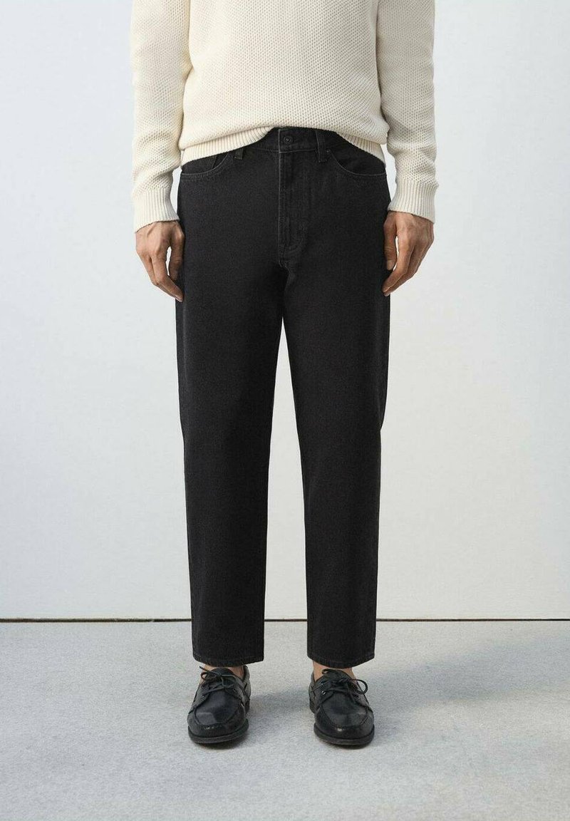 Osoba v krémovom pletenom svetri, čiernych nohaviciach s rovnými nohavicami a čiernych šnurovacích topánkach stojaca na svetlosivom podlahe pred bielou stenou.