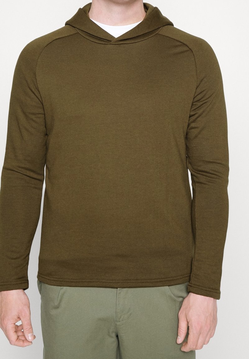 Homme portant une chemise à capuche à manches longues vert olive et un pantalon vert olive clair, debout devant un fond uni clair.