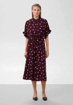 Femme portant une robe midi bordeaux à pois blancs, manches courtes, taille nouée, et chaussures plates noires, debout devant un fond uni.