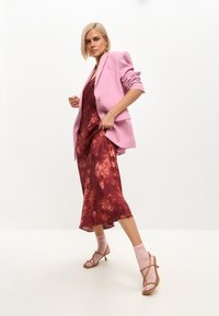 Jachetă roz deschis peste o rochie de slip roșie cu flori, asortată cu șosete roz și sandale cu toc bej. Texturi netede și un contrast îndrăzneț de culori.