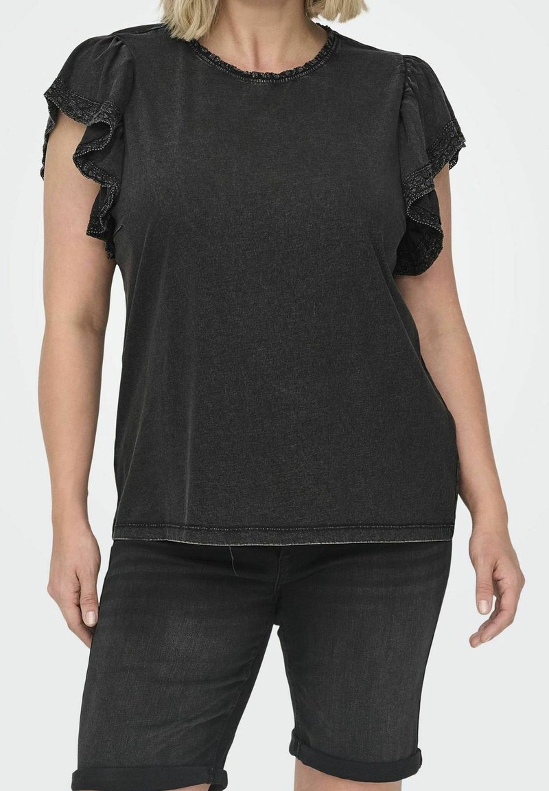 Femme portant un haut noir à manches courtes avec des bords volantés et un short en jean noir retroussé, debout devant un fond uni.