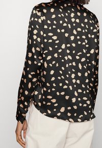 Camicia nera in satin con un motivo beige a pois, maniche lunghe, orlo arrotondato e una texture liscia. Un leggero lucido mette in risalto il design.