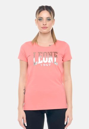 Giovane donna con due code di cavallo che indossa una maglietta rosa corallo "LEONE 1947" e pantaloni neri, in piedi davanti a uno sfondo bianco semplice.