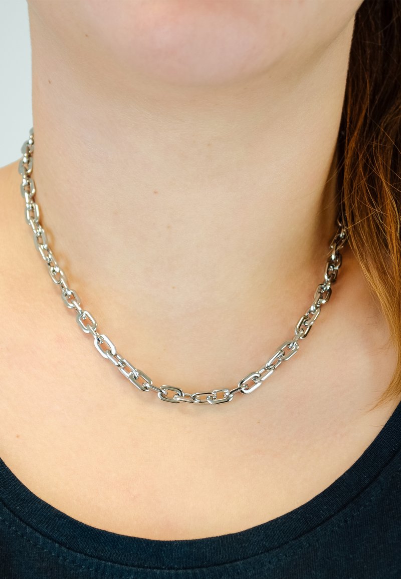 Zilveren ketting met langwerpige ovale schakels, gepolijste afwerking, comfortabel om de nek zittend, tegen een donkere shirt achtergrond.