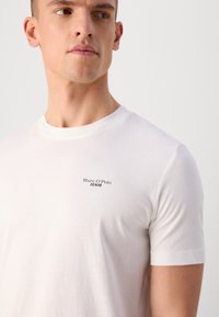 Bijela pamučna majica s okruglim izrezom, kratkim rukavima i malim crnim logom "Marc O'Polo DENIM" na prsima, glatke teksture.