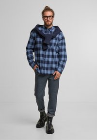 Cămașă de flanel cu carouri albastre și bleumarin, design cu butoni, asortată cu pantaloni din țesătură gri și botine negre până la gleznă. Pulover aruncat pe umeri.