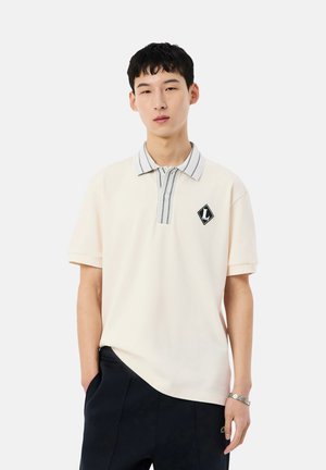 Joven con polo crema con cuello a rayas y parche de logo negro, pantalones negros y reloj de pulsera plateado sobre fondo blanco.