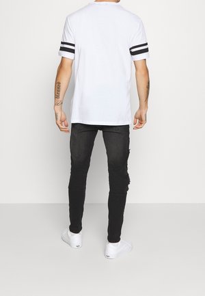 Camiseta blanca de manga corta con rayas negras en las mangas, combinada con jeans negros desgastados y zapatillas blancas.