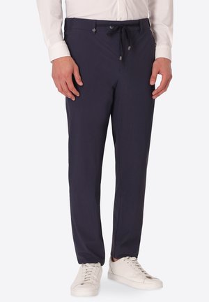 Uomo con pantaloni blu scuro con coulisse, camicia bianca a maniche lunghe e sneakers bianche, in piedi davanti a uno sfondo semplice.