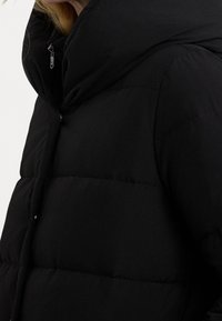 Černá vycpaná bunda s vysokým límcem, horizontálním prošíván ím a předním zipem. Materiál vypadá hladce a matně.