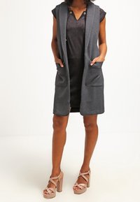 Graue ärmellose Weste mit langer, offener Vorderseite und zwei vorderen Taschen, kombiniert mit einem kurzen schwarzen Kleid und beigen High-Heels.