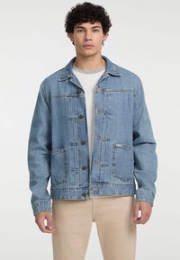 Chaqueta de mezclilla azul con cuello clásico, frente abotonado, dos bolsillos delanteros y detalles de costura. Lleva puesta una camiseta blanca.