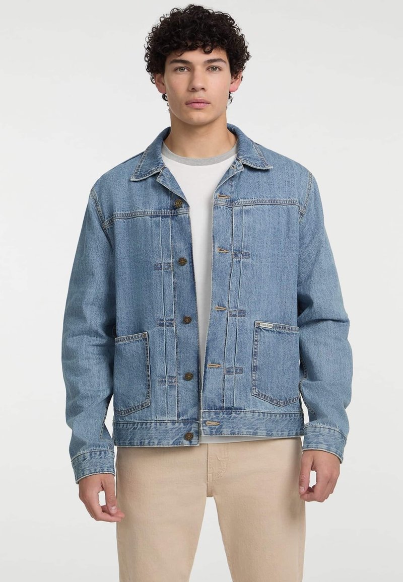 Chaqueta de mezclilla azul con cuello clásico, frente abotonado, dos bolsillos delanteros y detalles de costura. Lleva puesta una camiseta blanca.