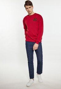 Felpa rossa con scollatura rotonda e dettaglio del simbolo ricamato sul petto, abbinata a jeans scuri e sneakers bianche.
