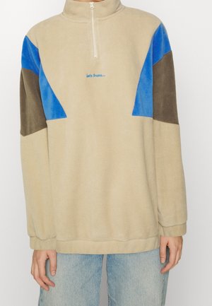 Pull polaire beige avec demi-fermeture éclair, panneaux bleu et marron aux épaules, et petit texte bleu sur la poitrine, porté avec un jean bleu clair.