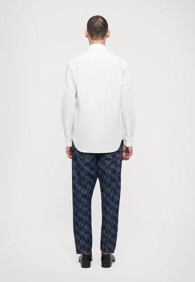 Camisa blanca de manga larga con un dobladillo redondeado, combinada con pantalones azul oscuro que presentan un patrón de logotipos repetidos. Zapatos negros completan el atuendo.