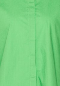Grünes Stoffhemd mit glatter Textur, geradem Schnitt und einer vorderen Nahtdetails. Die Farbe ist lebhaft und einheitlich.