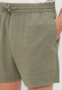 Κοντινό πλάνο ενός ατόμου που φορά σορτς με κορδόνι σε ελιά πράσινο χρώμα, με το χέρι στην δεξιά τσέπη, σε λευκό φόντο.