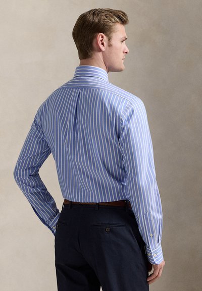 Camisa de homem às riscas azuis e brancas, de manga comprida, ajuste personalizado, punhos com botões, com um prega nas costas e textura leve. Usada com calças escuras.