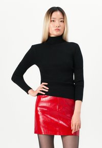 Νεαρή γυναίκα με μακριά ξανθά μαλλιά, φορώντας μαύρο πουλόβερ τύπου τζόκερ, γυαλιστερή κόκκινη δερμάτινη μίνι φούστα και μαύρες κολάν, στέκεται με το χέρι στη μέση.