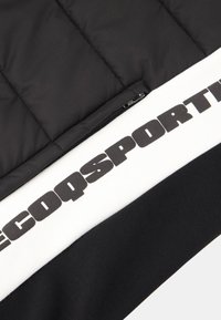 Doudoune noire avec une surface matelassée, comportant une bande blanche avec un texte noir en gras et une poche zippée sur le côté.