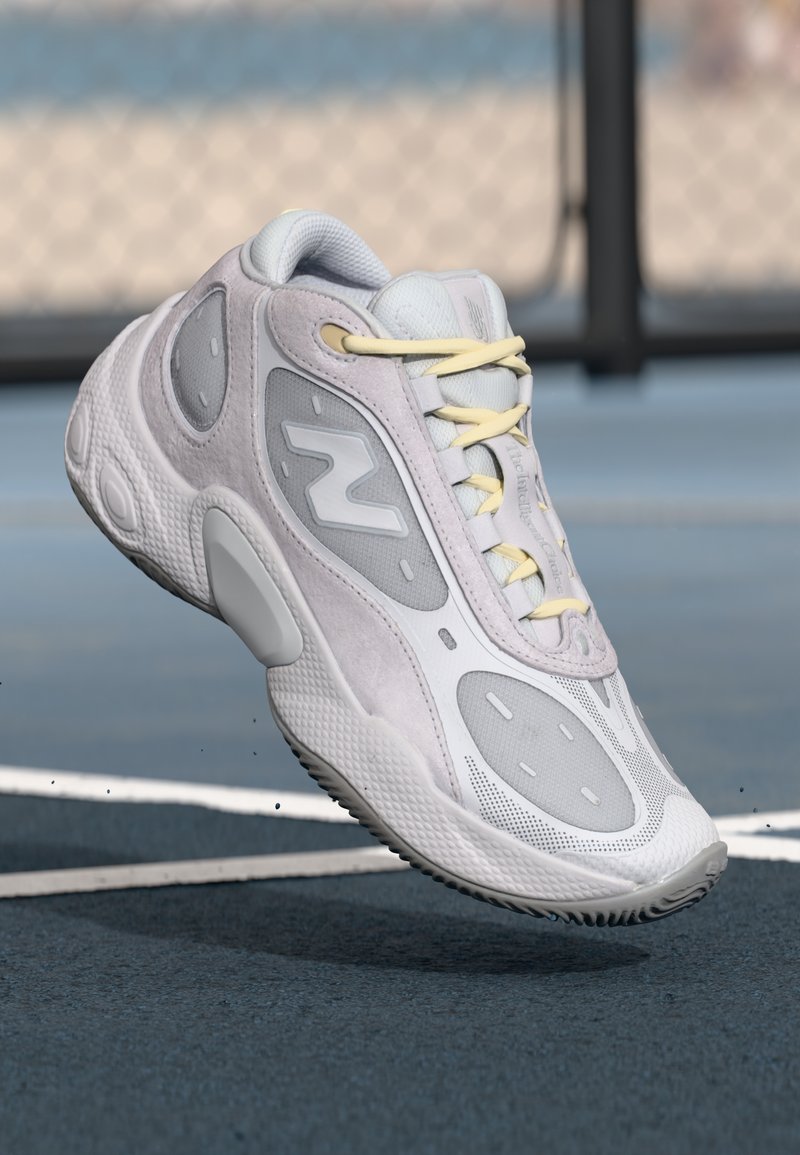 Hellegraue Sportschuh mit einem strukturierten Mesh-Obermaterial, weichen Wildlederakzenten, gelben Schnürsenkeln und einem markanten N-Logo an der Seite.