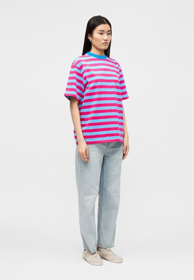 Νέα γυναίκα στέκεται, φοράει μπλε και ροζ ριγέ μπλουζάκι, ανοιχτό μπλε τζιν και μπεζ αθλητικά παπούτσια μπροστά σε απλό ανοιχτό φόντο.