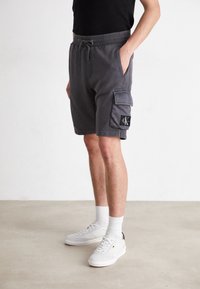 Shorts cargo gris avec une taille élastique et un cordon de serrage, dotés de poches latérales et d'une patch logo noir sur la jambe. Associés à des baskets blanches.