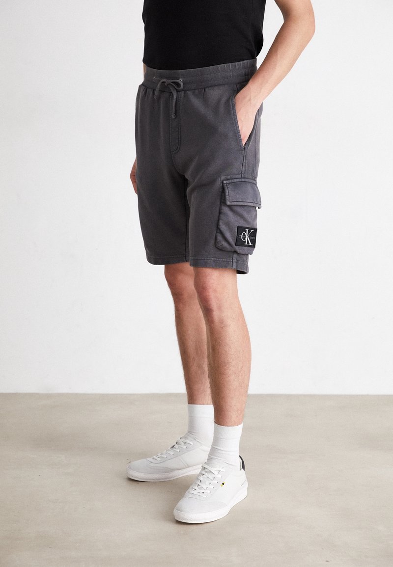 Shorts cargo gris avec une taille élastique et un cordon de serrage, dotés de poches latérales et d'une patch logo noir sur la jambe. Associés à des baskets blanches.