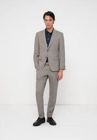 Ανοιχτό γκρι κοστούμι από υφασμάτινη υφή, με σακάκι ενός κουμπιού, κομψή εφαρμογή και μαύρα παπούτσια. Από κάτω, ένα σκούρο πουκάμισο.