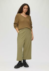 Bruine, gestructureerde V-hals trui gecombineerd met op maat gemaakte, losvallende olijfgroene broek. Zwarte veterschoenen tot de enkel maken de outfit compleet.