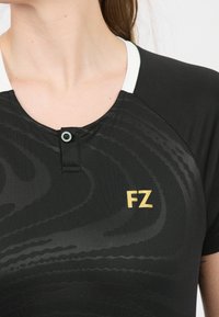 Svart atletisk tröja med texturerat mönster, vit krage och knappstängning. Har en gyllene "FZ"-logotyp på framsidan.