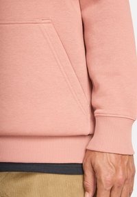 Close-up of a person wearing a pink sweatshirt with a front pocket, ribbed cuffs, and beige corduroy pants.